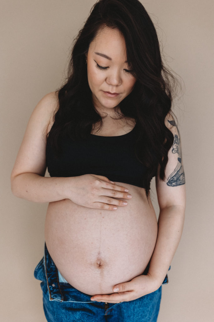 Pregnant woman standing with hands on her belly in a studio while wearing a black bra and jeans.