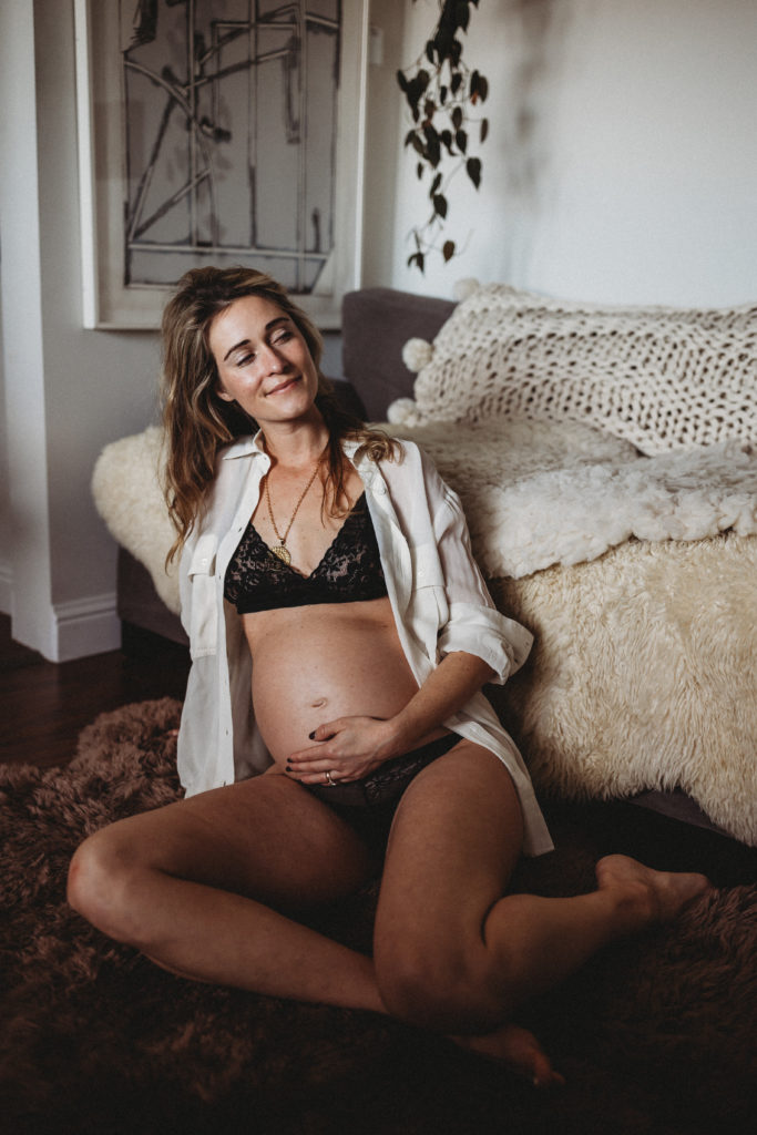 Pregnant woman sitting on floor in front of couch in underwear and white button up shirt with hand on belly.