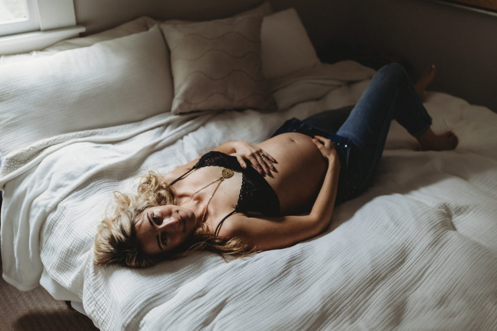 pregnant woman laying on bed in bra and jeans with hands on belly and looking at the camera, intimate maternity photo, pregnancy photo