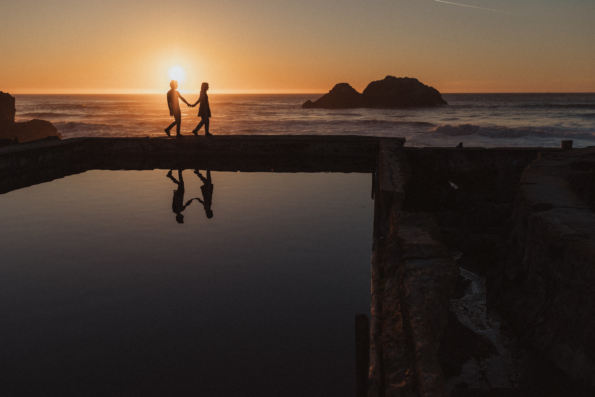 Boy and girl holding hands and walking across Sutro Baths at sunset for their engagement photo session in San Francisco.