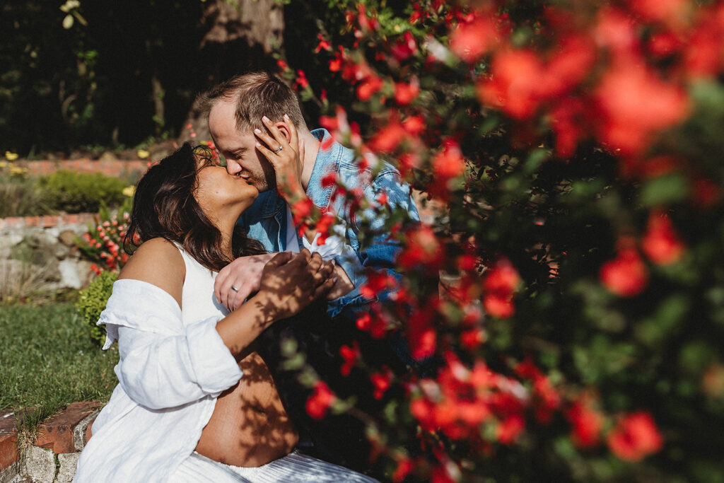 Expecting parents kissing with lush flowers in foreground of image.