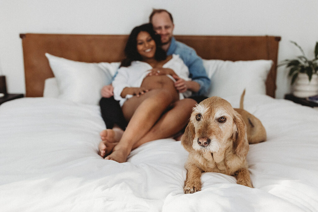 Expecting parents lounging on their bed at home with their dog at their feet.