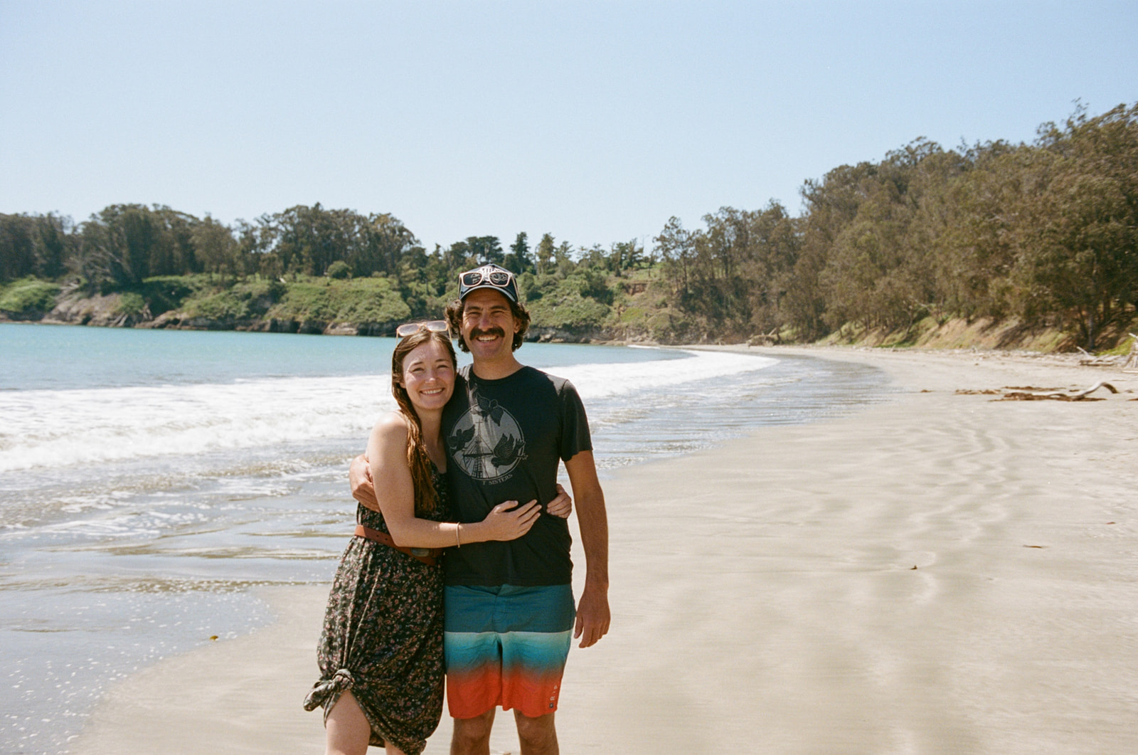 Man and wife standing together on a beach in Cambria, California and smiling at the camera.