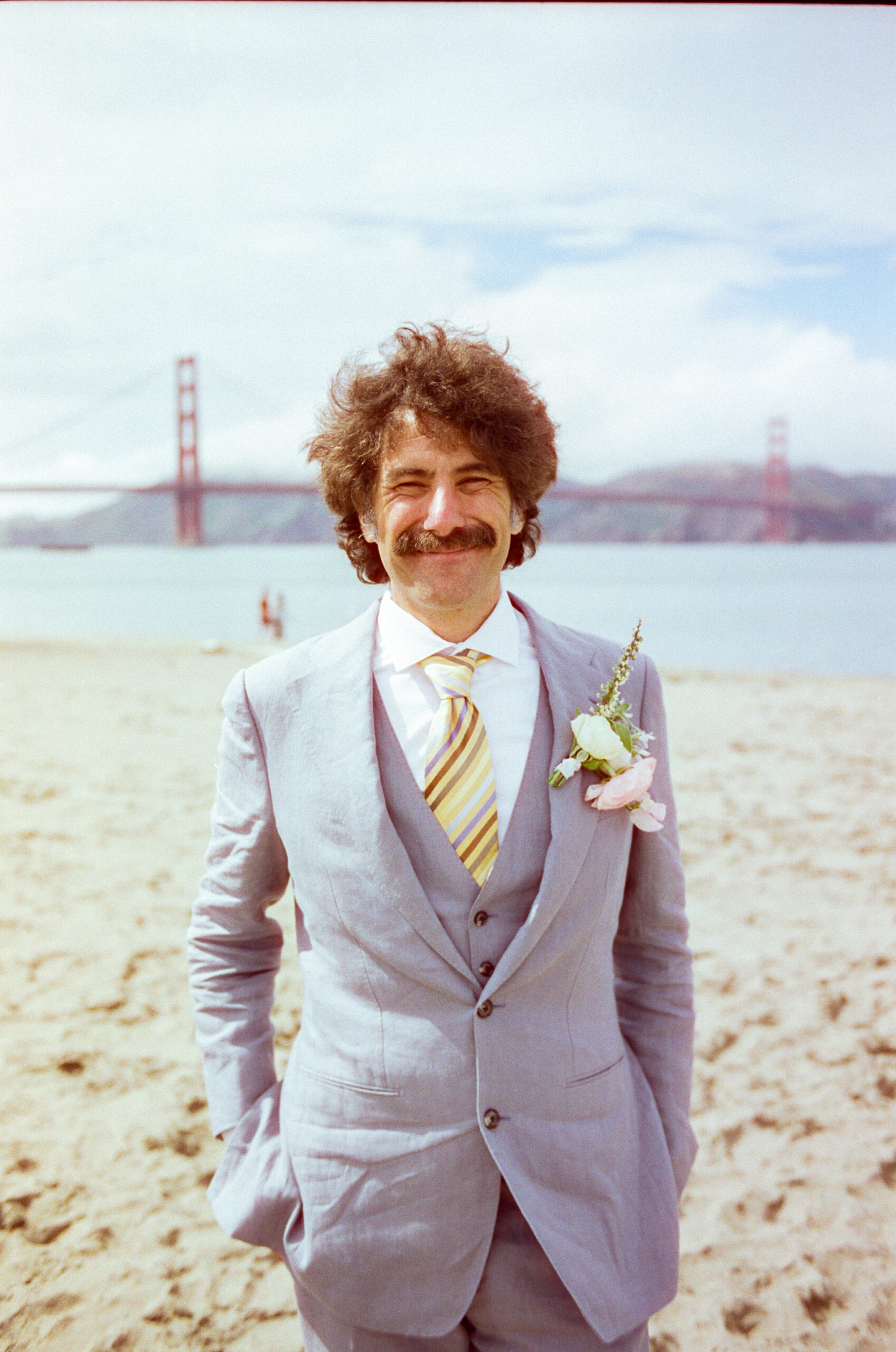 Medium close up of a groom standing on Crissy Field Beach with his hands in his pockets and the Golden Gate Bridge behind him. He's smiling at his wife behind the camera.