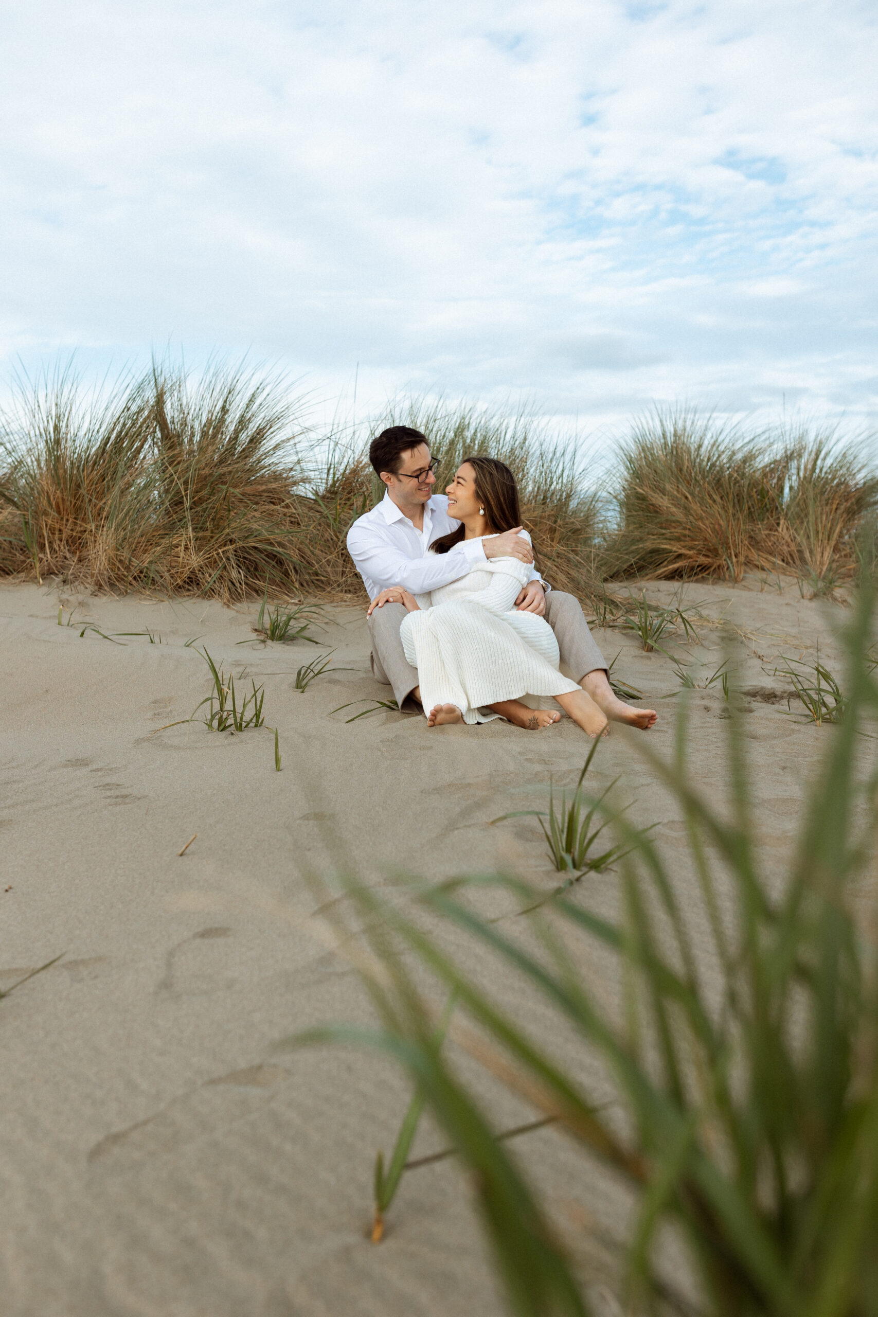 Husband and pregnant wife cuddled together while sitting on a sand dune at Ocean Beach in San Francisco, taken by Skyler Maire Photography.