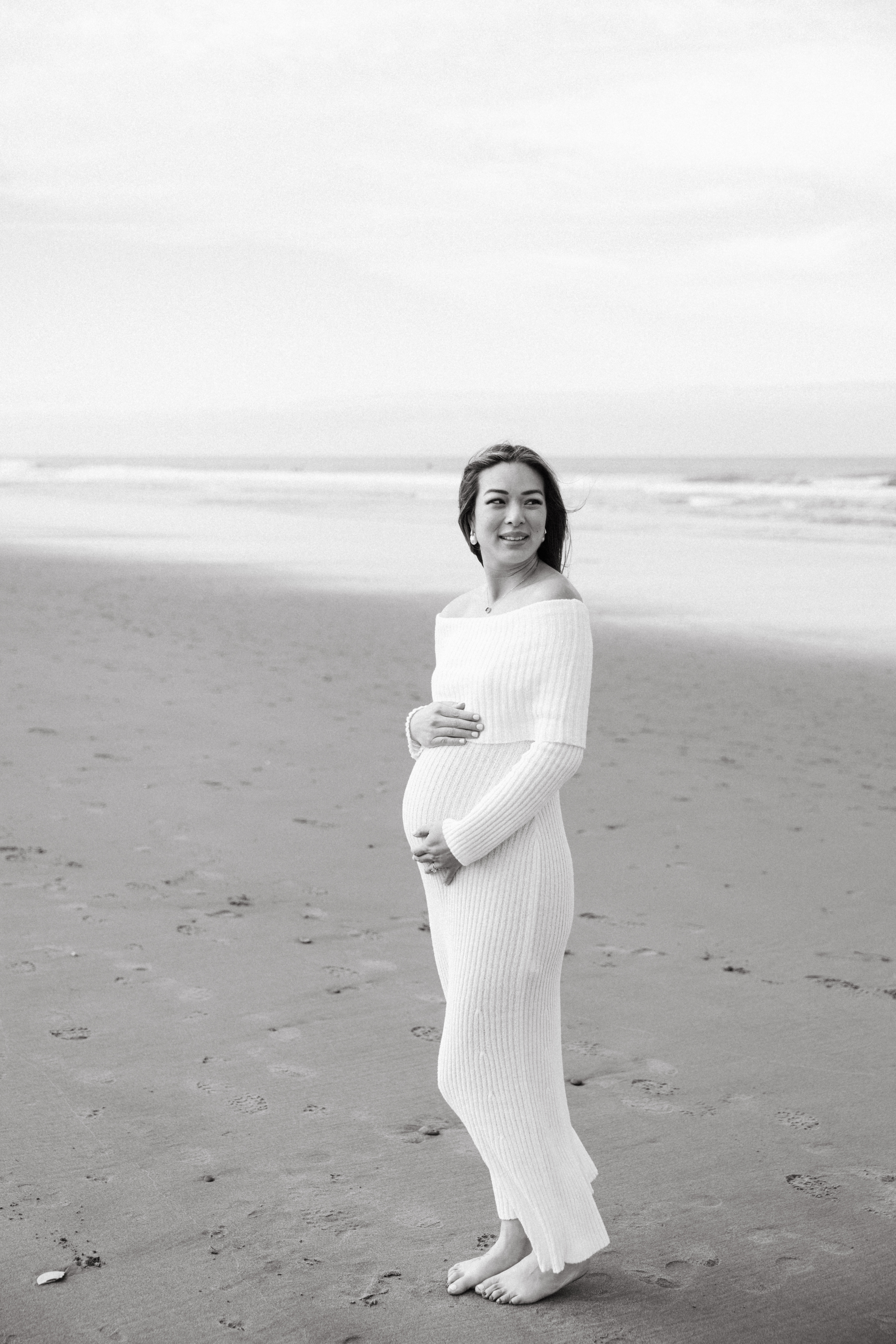 Black and white photo of a pregnant woman standing on Ocean Beach wearing a long sleeved white sweater dress and holding her belly. Taken by Skyler Maire Photography.