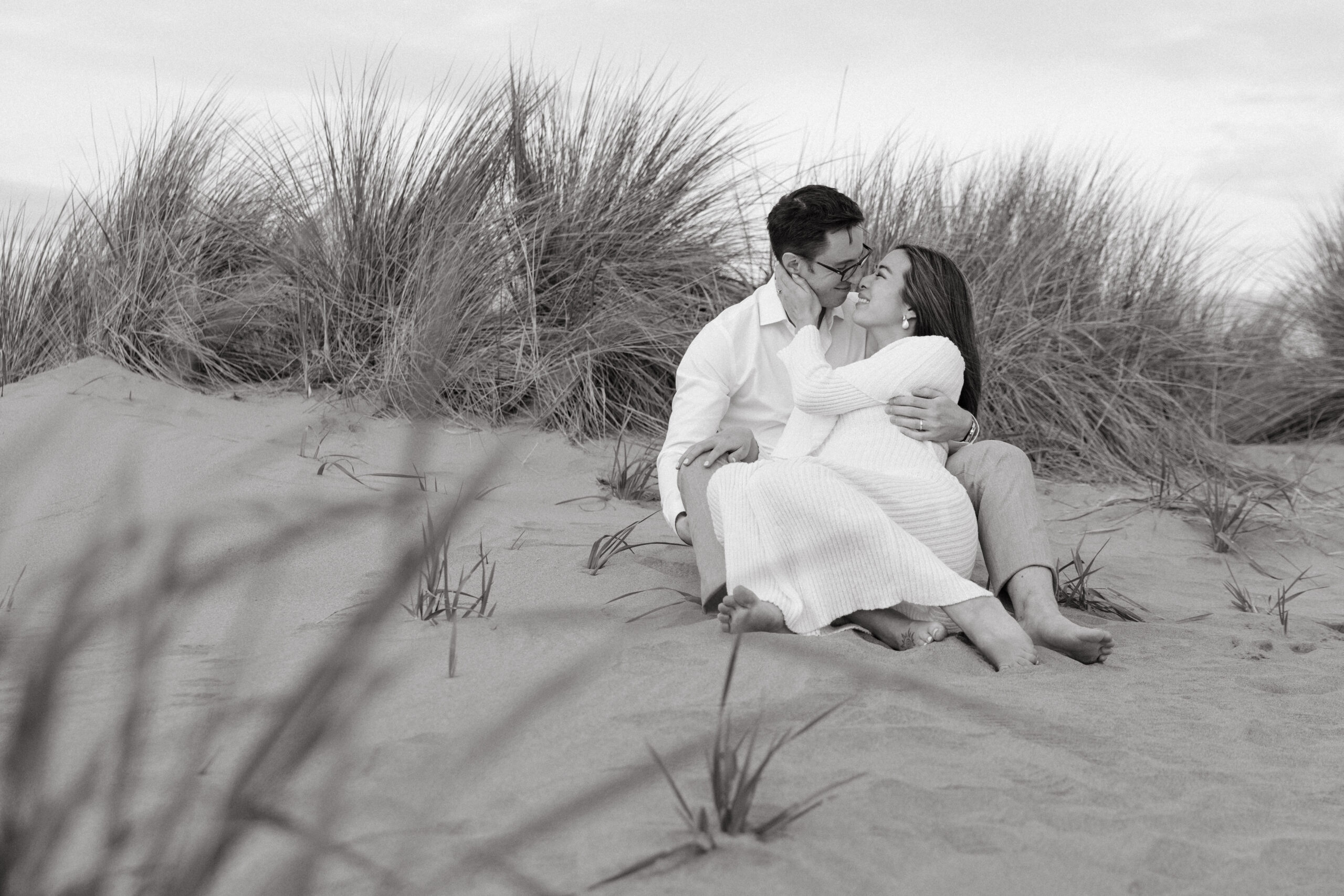 Husband and pregnant wife sitting on a sand dune at Ocean Beach, San Francisco, and looking at each other while touching noses. Photo taken by Skyler Maire Photography.