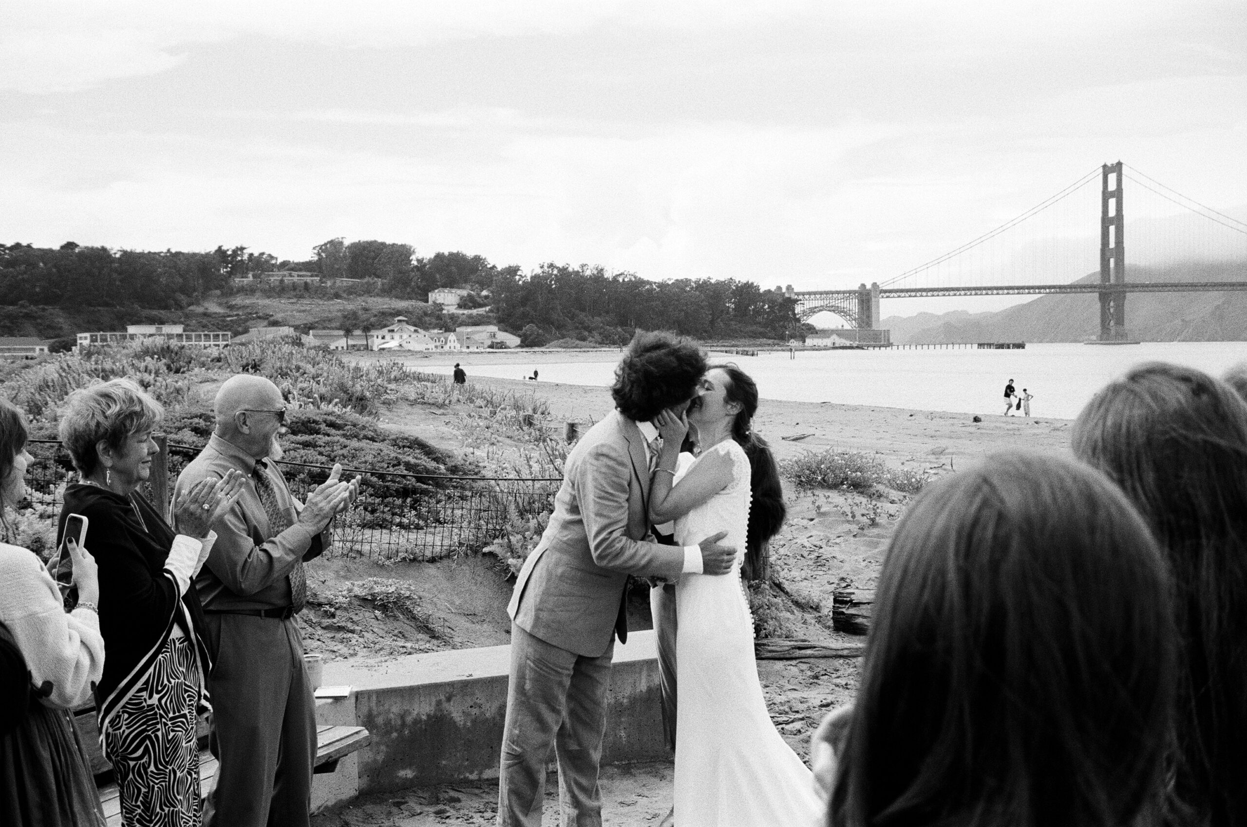 A couple kisses on a wooden platform on Crissy Field Beach in San Francisco during their wedding ceremony while their family claps next to them. The Golden Gate Bridge is in the distance.