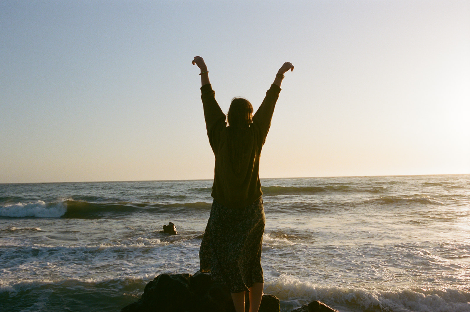 Silhouette of a girl standing on Moonstone Beach in Cambria and facing the water with her hands in the air and the sun setting in front of her.