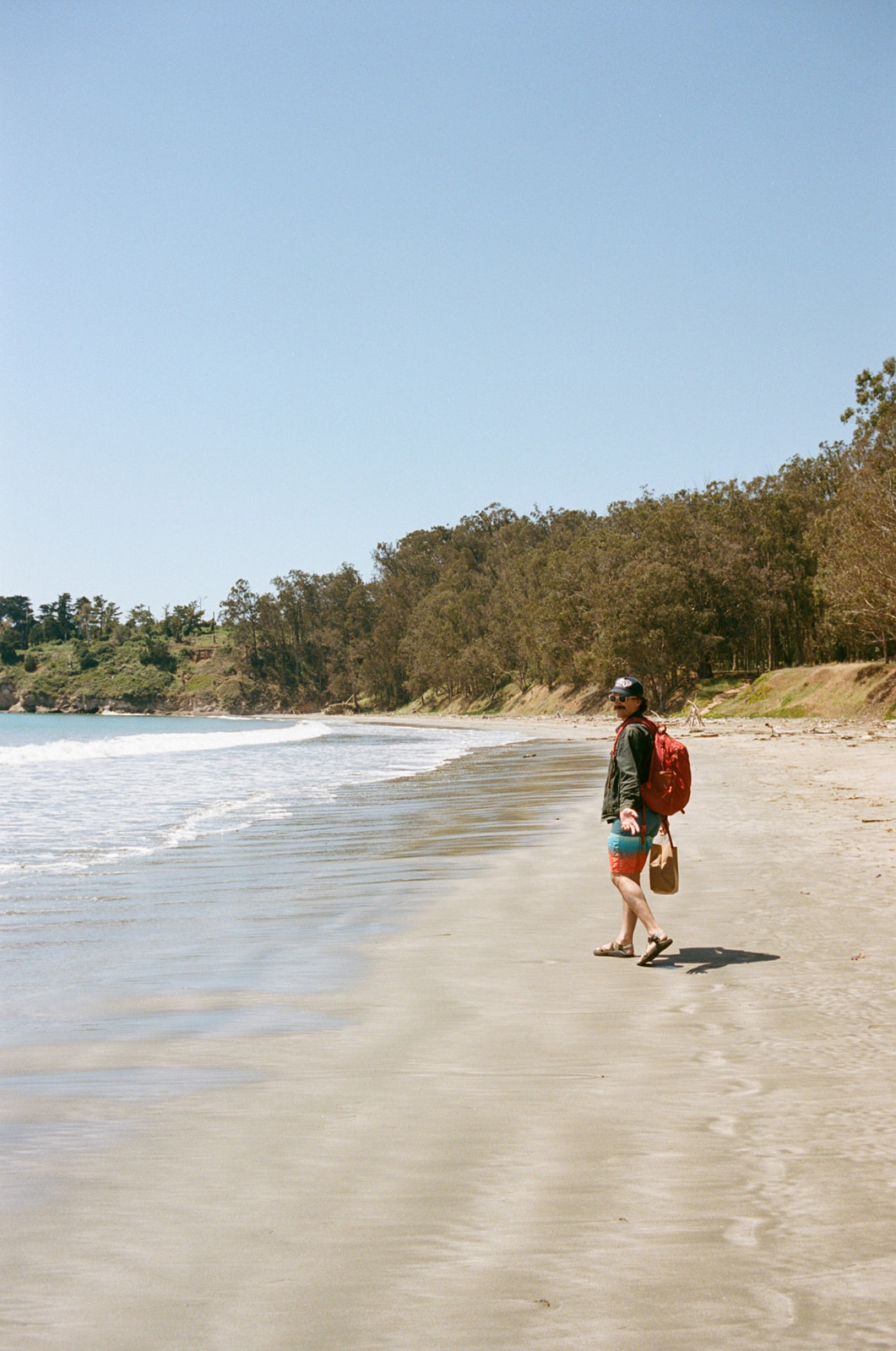 Wide photo of a man walking down a beach near Cambria, California, while wearing a red backpack. Taken by Skyler Maire Photography.