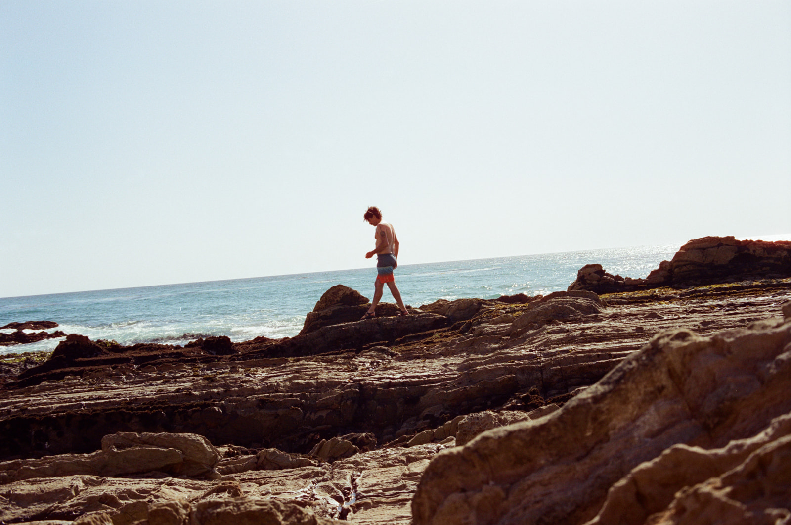 A wide photo of a man in swim trunks walking along a rocky coastline and looking at tide pools on the West Coast. Taken by Skyler Maire Photography.