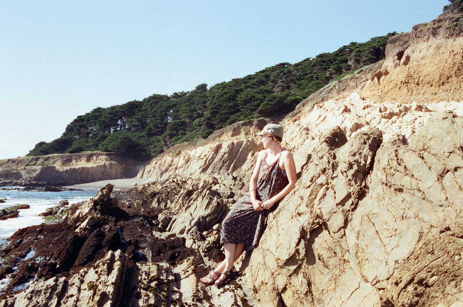 A girl leaning against rocks on a rocky coastline amongst tide pools, looking towards the ocean. 