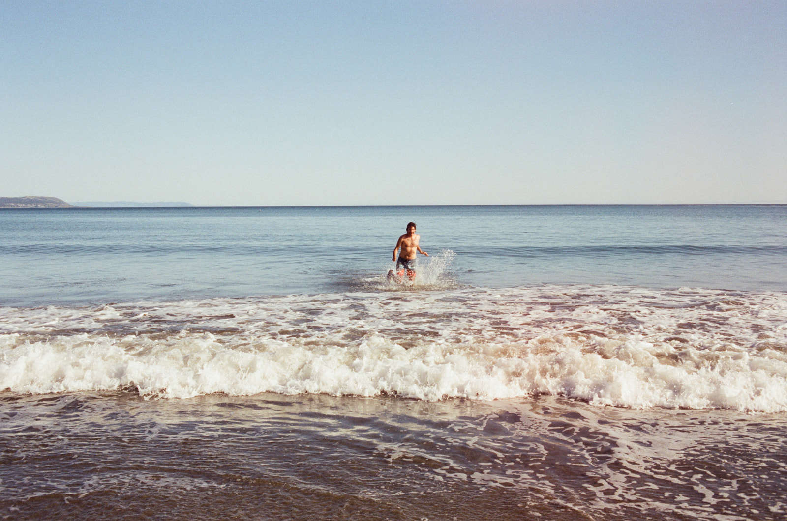 Happy man in the ocean running towards the beach and camera on the shore, taken by Skyler Maire Photography.