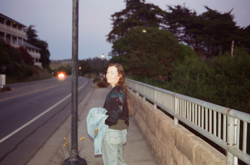 A woman walking down a sidewalk on the side of a road in Cambria, looking back at the camera at dusk.