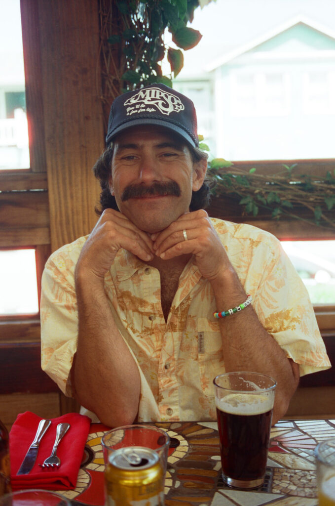 Cute man sitting at a lunch table and smiling at the camera while resting his chin on his hands. Taken by Skyler Maire Photography.