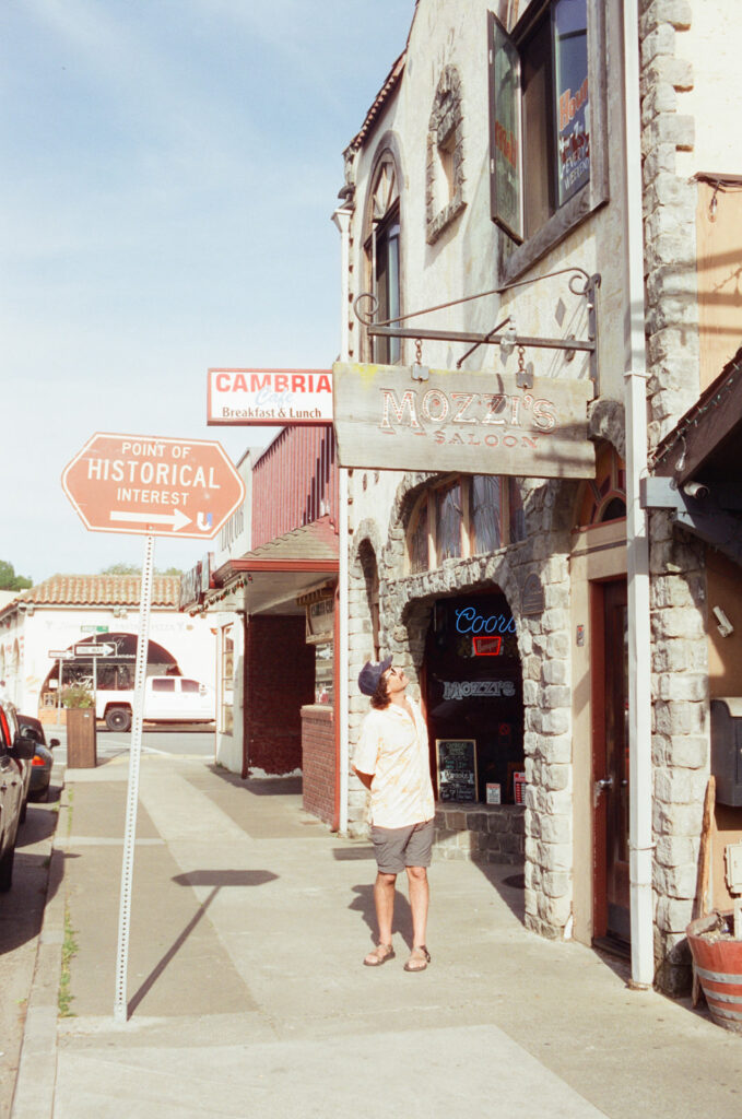 Man standing under Mozzi's Saloon sign in downtown Cambria, California. Taken by Skyler Maire Photography.