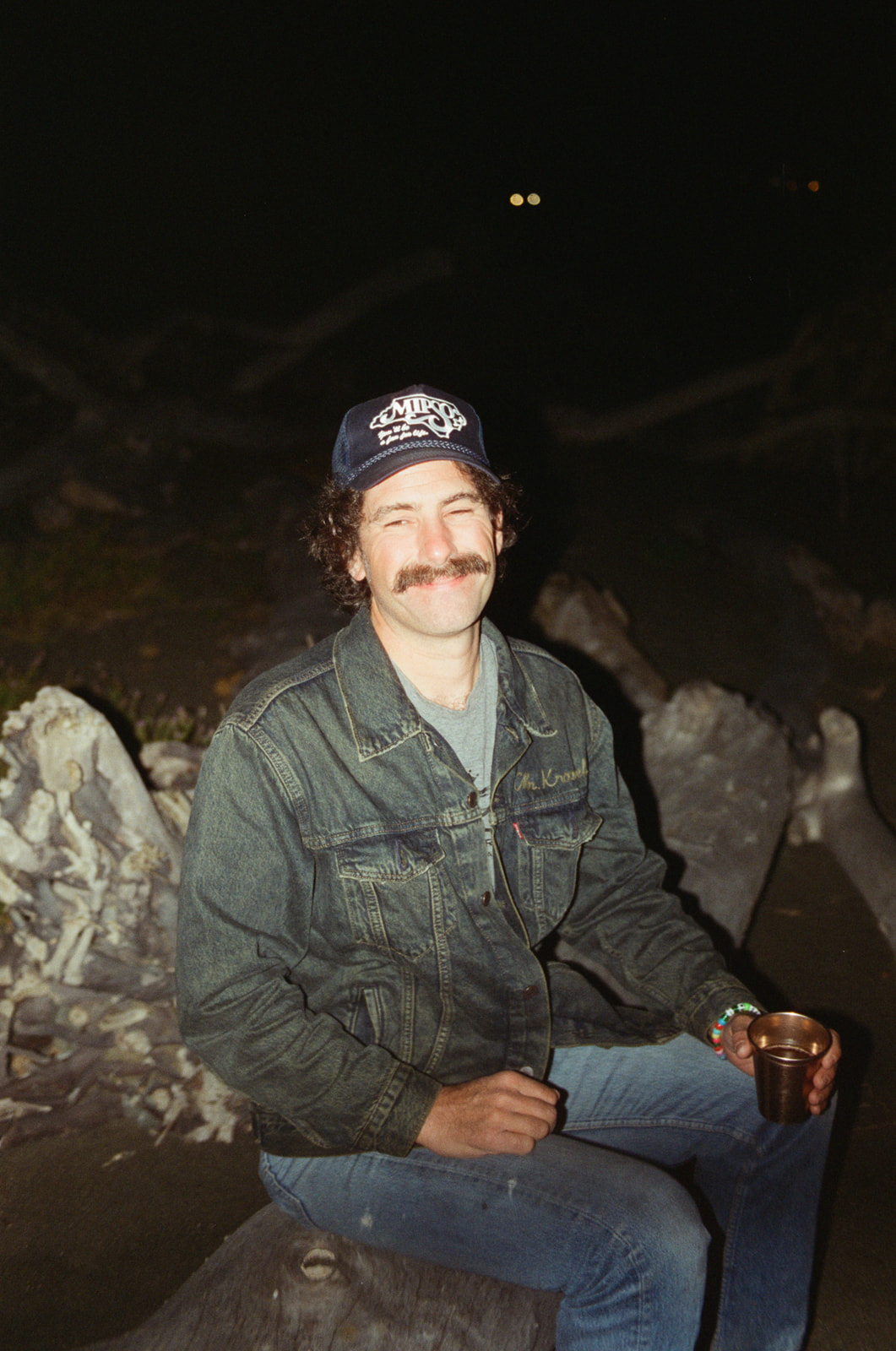 Man sitting on Moonstone Beach in Cambria after dark, drinking champagne out of a cup and smiling at the camera. Taken by Skyler Maire Photography.