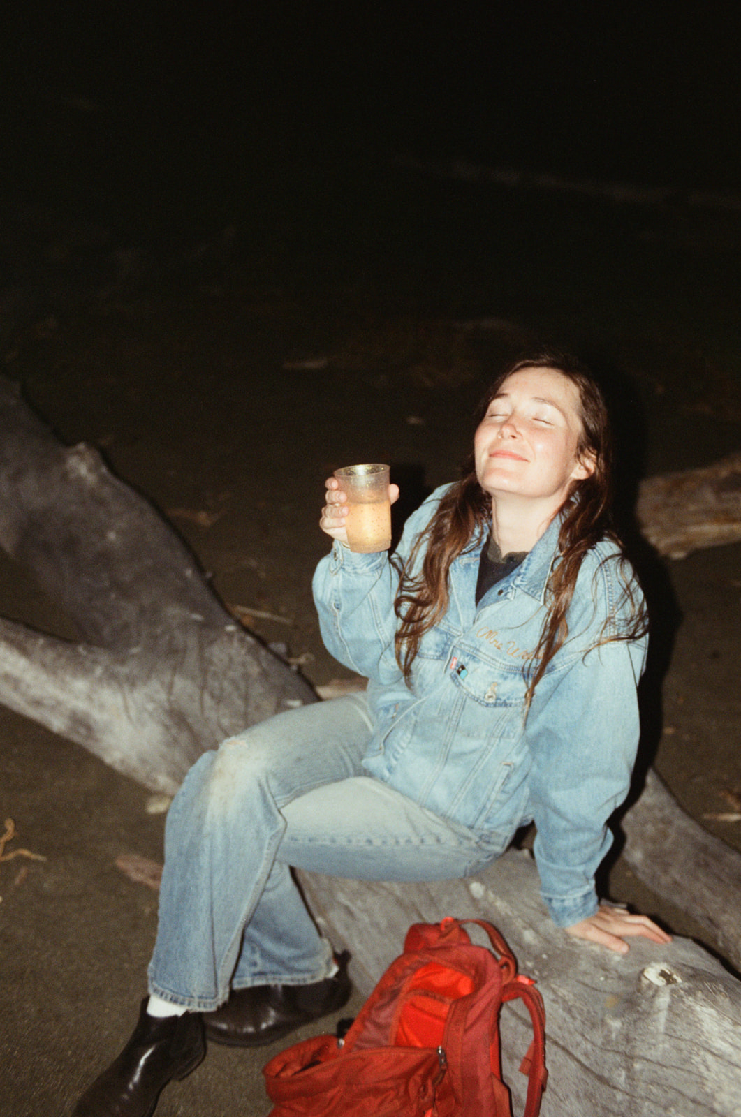 Girl sitting on a log on Moonstone Beach with a cup of champagne in her hand and smiling at the camera with eyes closed.