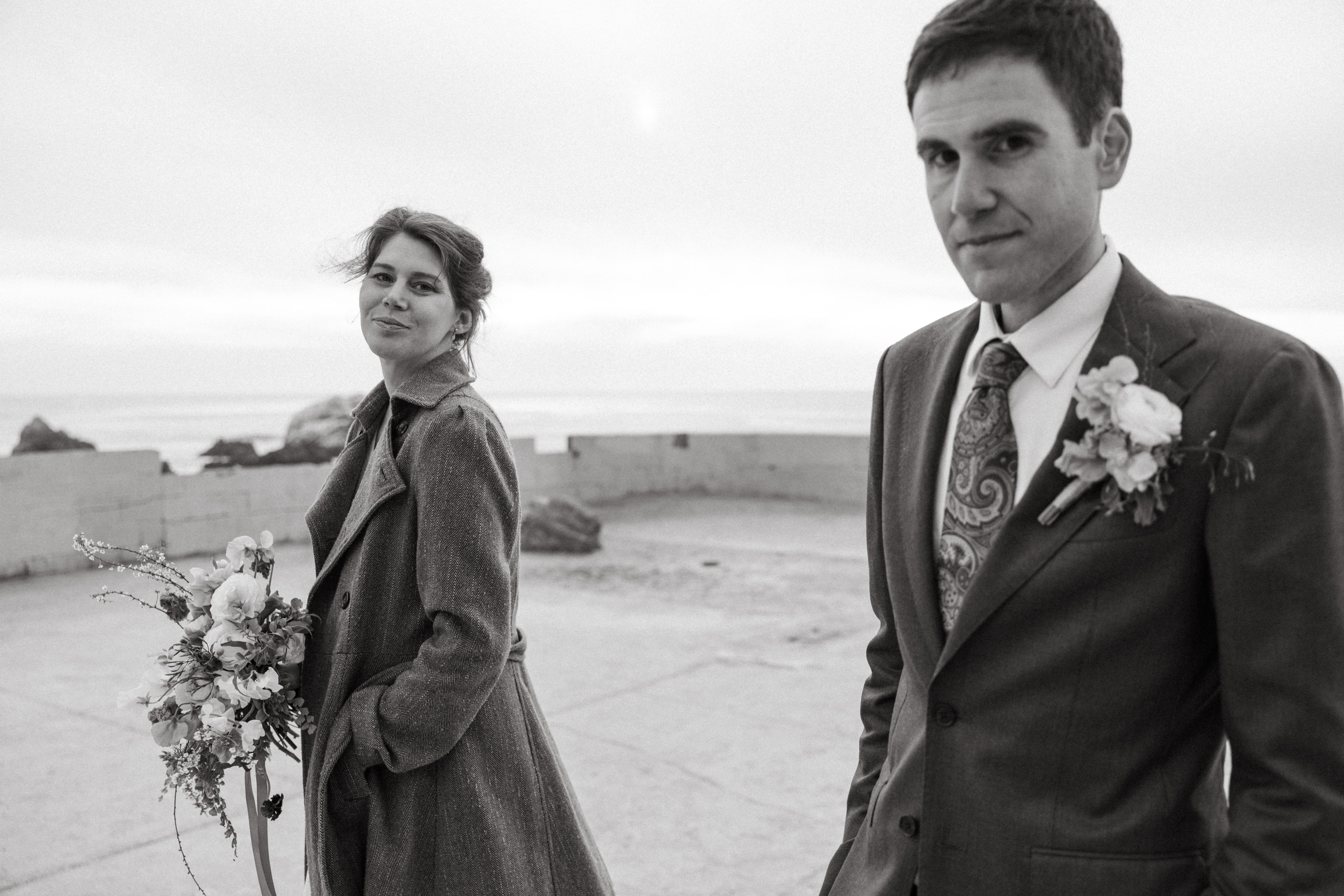 A black and white candid image of a bride and groom walking together and looking at the camera at San Francisco's Sutro Baths on a very foggy and overcast day.
