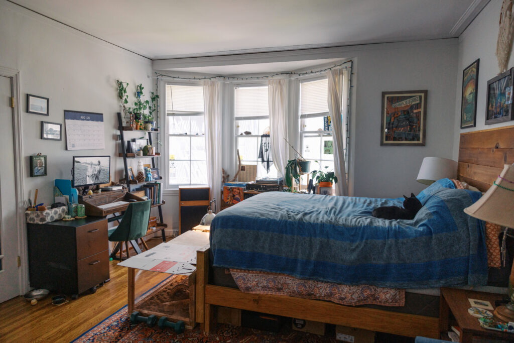 A small studio apartment living room in San Francisco with a bed, small desk at the foot of the bed, some shelving, and a record player in front of the bay windows in the back.