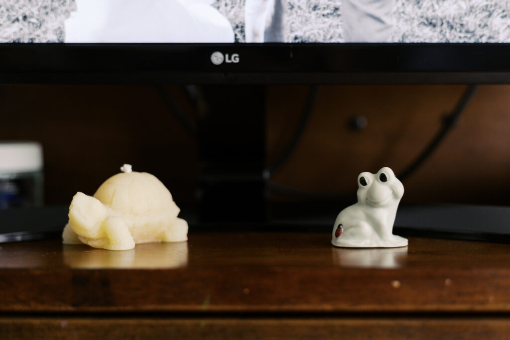 A small glass frog and candle in the shape of a tortoise in front of a computer monitor on a desk.